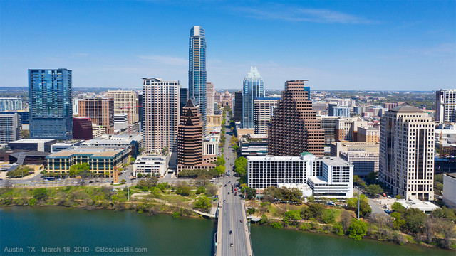 Cityscape of modest downtown with many tall buildings with many small appearing windows; photo is bisected by an avenue that seems to disappear in the distance at a domed capitol building; along the bottom a blue/green river which the avenue crosses; blue sky with a few thin clouds.
©BosqueBill.com