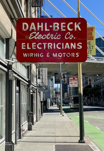 Photo looking down a sidewalk with building fronts on left and street on right. A red projection sign is directly ahead of the viewer. It has white letters in gothic and script styles. It reads: “DAHL-BECK Electric Co. Electricians Wiring & Motors”