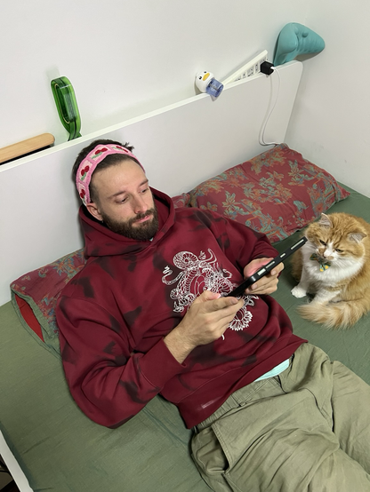 A man in a red sweatshirt lies on a bed reading from an iPad. Next to him sits an orange and white cat and there is a hair band on his head