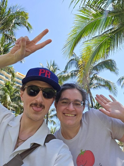 A mustachioed man in sunglasses and a PB hat sticking out his tongue and giving a peace sign, next to a waving brunette woman whose t-shirt has an illustration of a tomato drinking tea and says “toma-té”