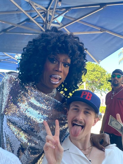 A drag queen dresses in silver with black curly hair sings next to the friend with the mustache, who is giving a peace sign and has his tongue hanging out.