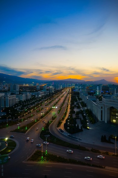 A colourful sunset over one of the major routes through the white marble city of Ashgabat. The signature white cars can be seen driving through the city lights and the mountains beyond meet the pastel sky.