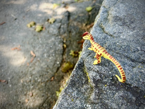 A green Dilophosaurus with brown spots stands on the edge of a stone wall and looks out at the sidewalk below. Fallen leaves and maple keys dot the concrete path beneath them.
