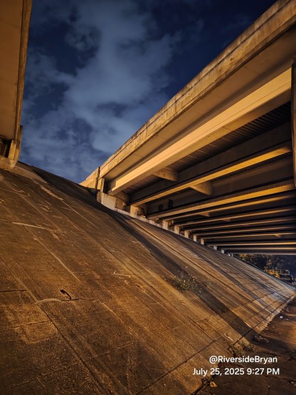 Late night view up into the deep blue sky with smoke-like clouds in the gap between the overhead lanes of a highway bridge overpass, illuminated at night showing age, weathering and dirt.