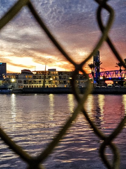 From behind a chain link fence looking out over a river where an explosion of colors overtake the sky, illuminating downtown skyline and bridges.