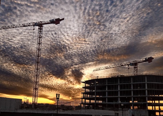 Intense sky-drama above a dark construction site, with silhouettes of tall work cranes and the skeleton of a new construction building against the sun down in a purple-grey night sky, blanketed with a sheet of cotton ball like clouds over the firey streaks of yellow, orange, and red at the horizon.
