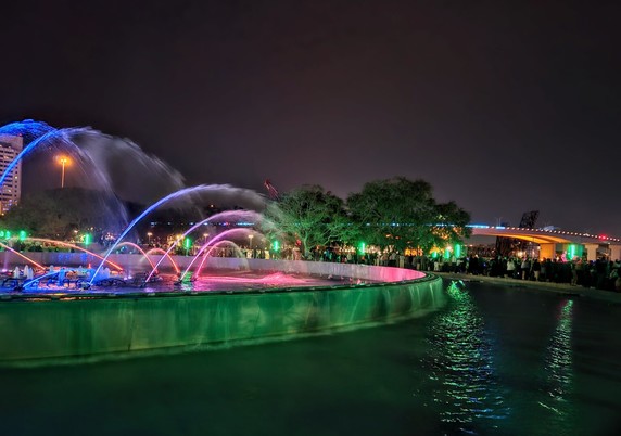 Late night lights brighten the surroundings of a view from a city park on the riverfront overlooking a colorful bridge and downtown skyline casting reflections upon the river below while in the foreground a massive fountain with jets of water illuminated with a variety of colors reflects onto a pool below.