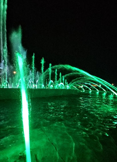 Green lights illuminate a large fountain with water jet streams against dark black night sky.