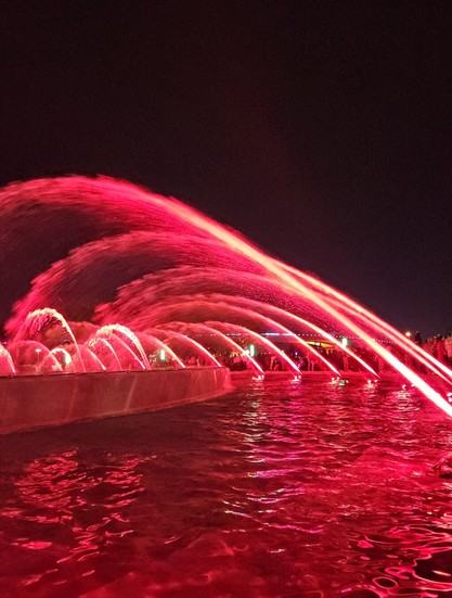 Red lights illuminate a large fountain with water jet streams against dark black night sky.