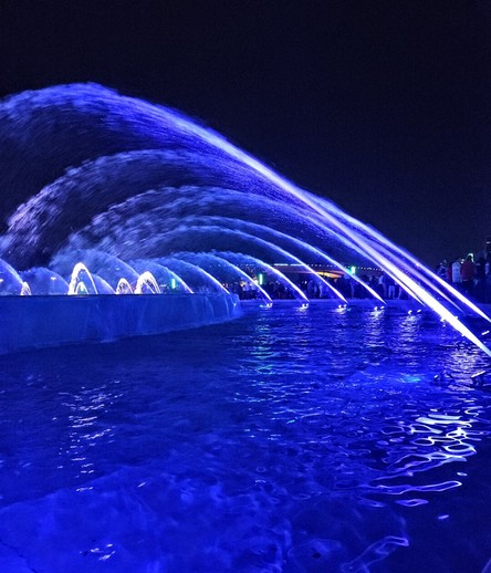 Blue lights illuminate a large fountain with water jet streams against dark black night sky.