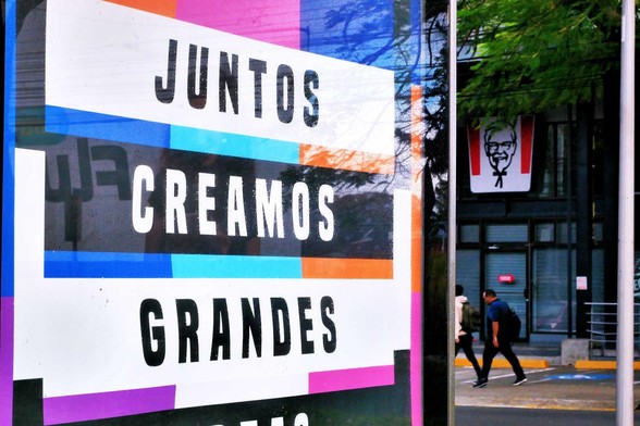 Foreground: Large sign with the phrase "Juntos creamos grandes"; background: two men walking in front of a KFC restaurant