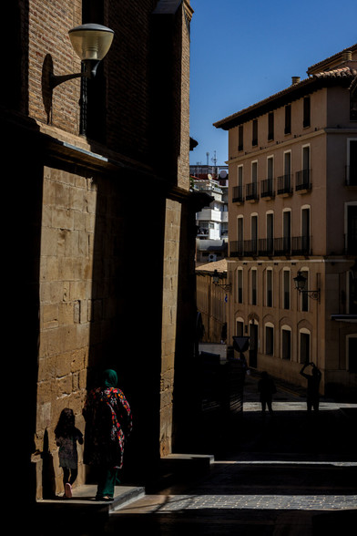 Narrow street with strong contrasts of light and shadow. On the left, a woman wearing a green headscarf and a patterned coat walks with a small child close to a shaded wall. In the background, sunlit buildings and a deep blue sky.
