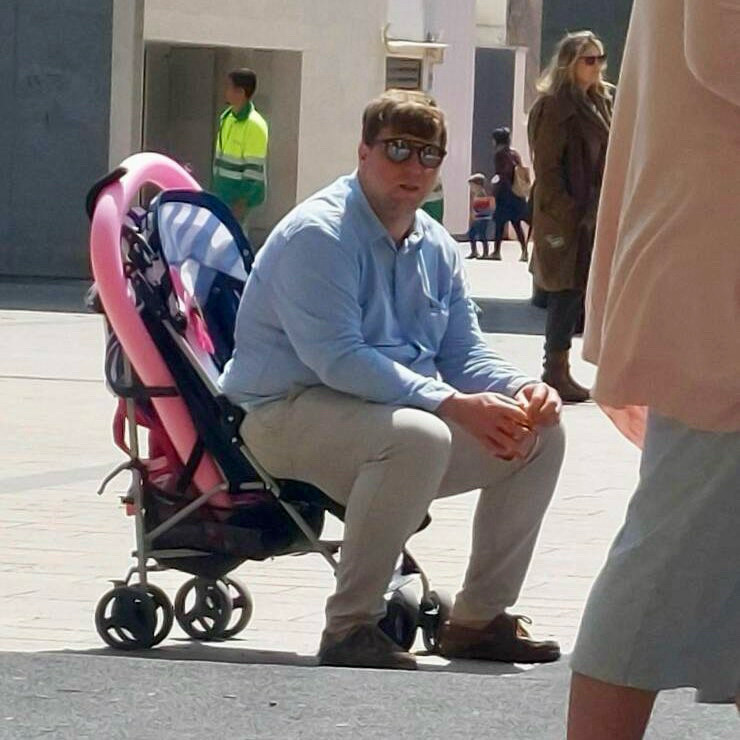 A man seated on a pink stroller, outdoors on a sunny day. Other people are visible in the background. A man seated on a pink stroller, outdoors on a sunny day. Other people are visible in the background.