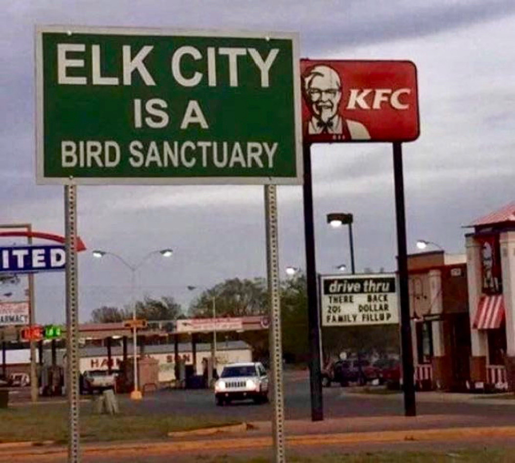 Road sign reading “ELK CITY IS A BIRD SANCTUARY” with a KFC sign behind and to its right.
