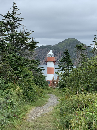 A narrow path leads through greenery towards a solitary lighthouse with red and white stripes, set against a backdrop of rocky hills and a cloudy sky.