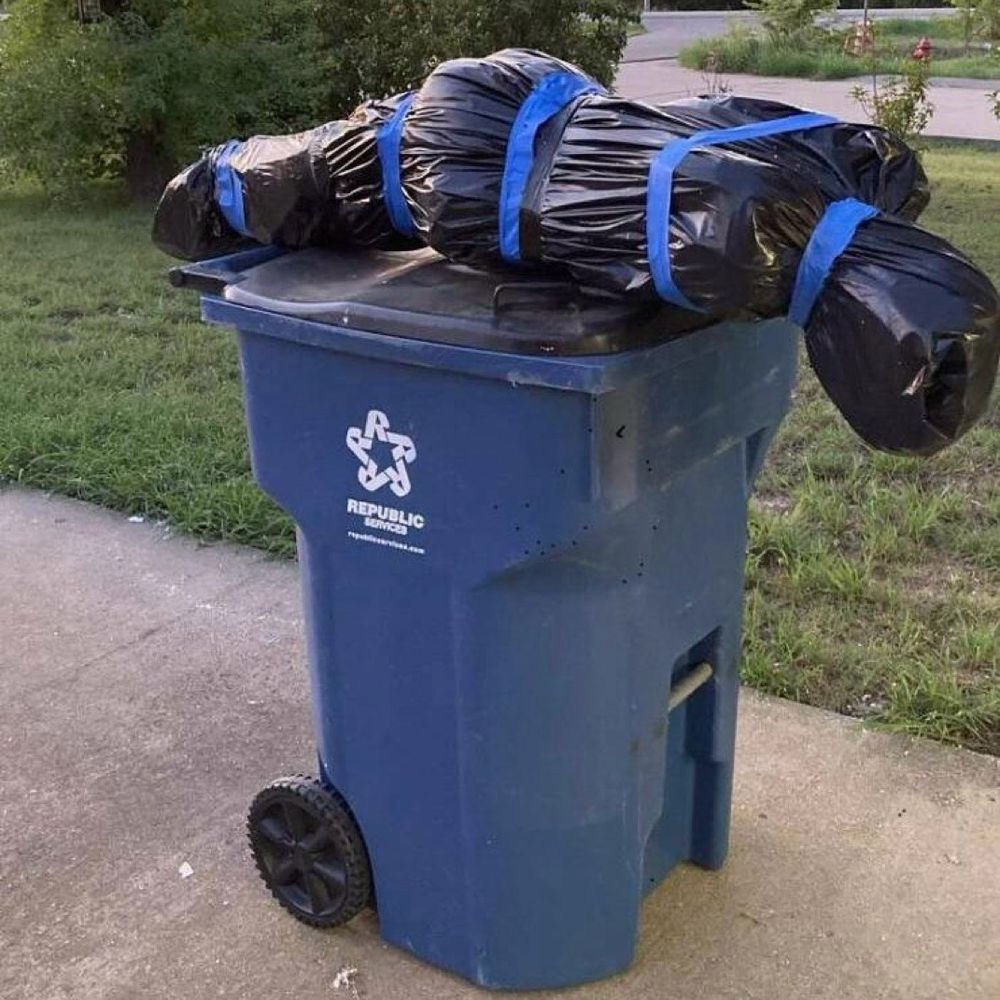 A body-shaped object wrapped in black plastic and painter’s tape sits atop a recycling can by the curb.