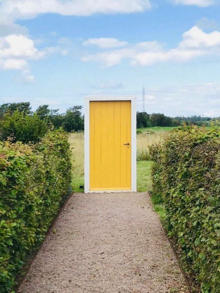 A framed yellow door sits at the end of a path between two rows of hedges. Behind the door is an open meadow. A framed yellow door sits at the end of a path between two rows of hedges. Behind the door is an open meadow.